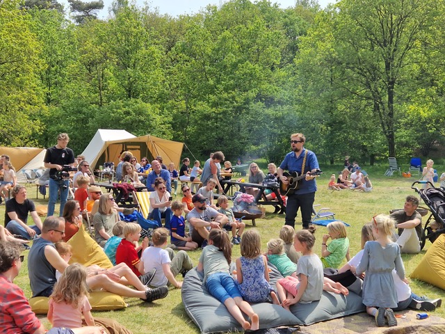 Een man speelt gitaar in de openlucht. Om hem heen zitten kinderen en volwassenen in een grote kring op de grond.